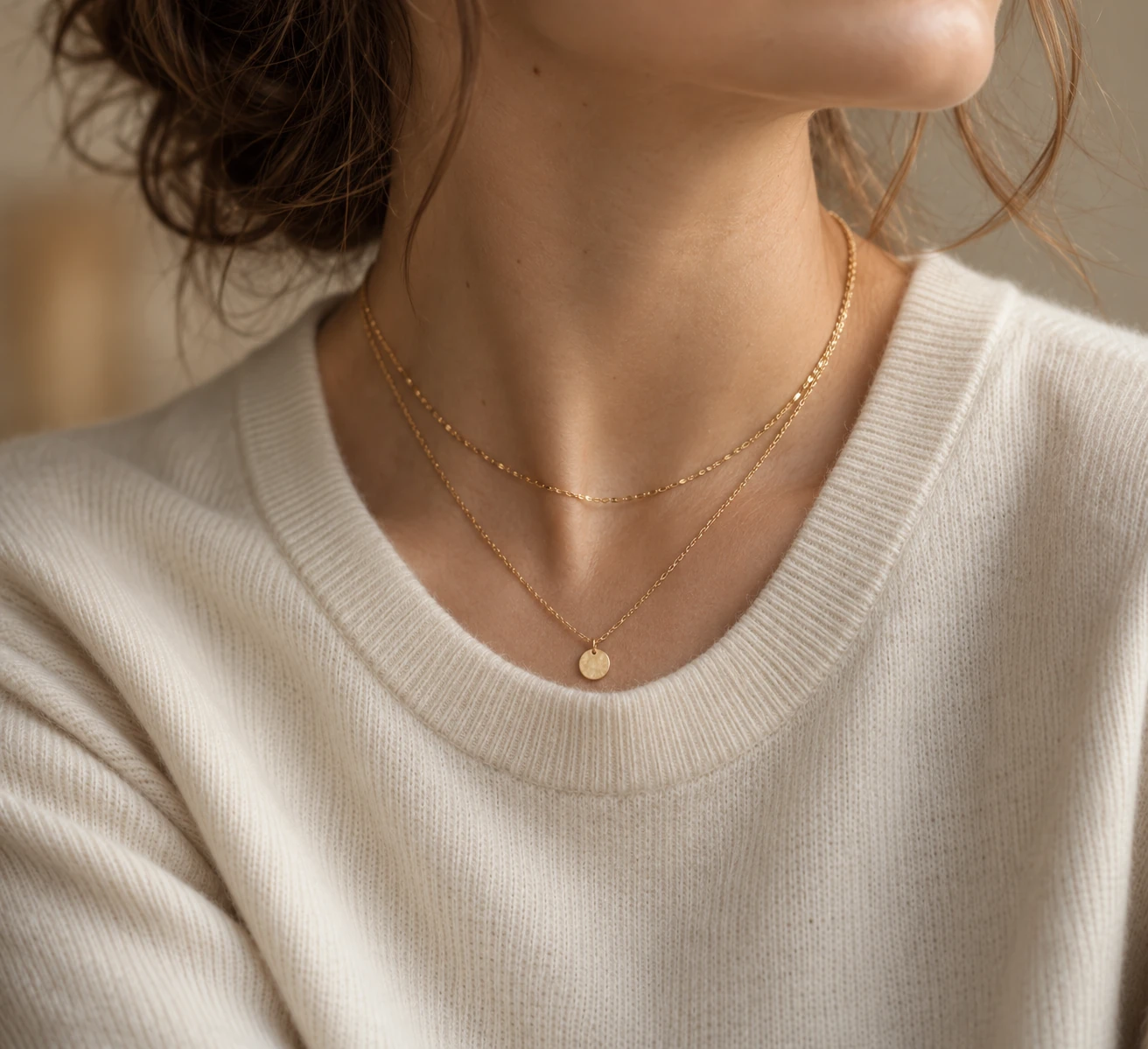 Close crop of a cream crew-neck knit worn with two fine gold chains at clearly different lengths.